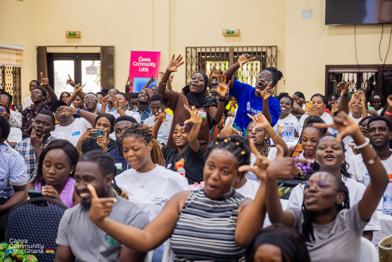 Rows of attendees on their feet with their arms raised, making C shapes with their hands and cheering towards the stage.